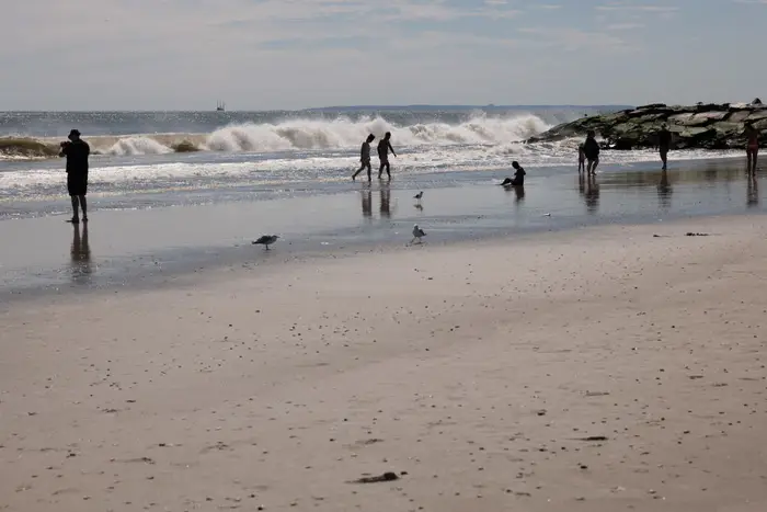 People walk along the water on Rockaway Beach as a wave breaks in the distance.